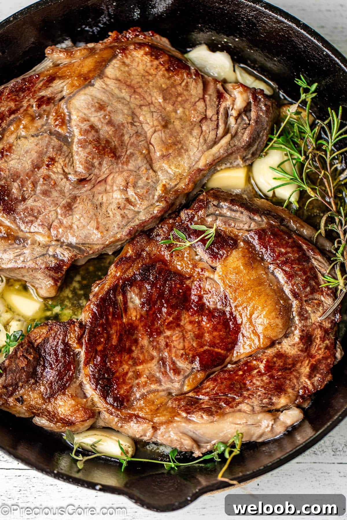 Perfect Pan-Seared Ribeye Steak with Crispy Crust and Juices 2 Two ribeye steaks in a cast iron skillet with rosemary, thyme, and garlic.