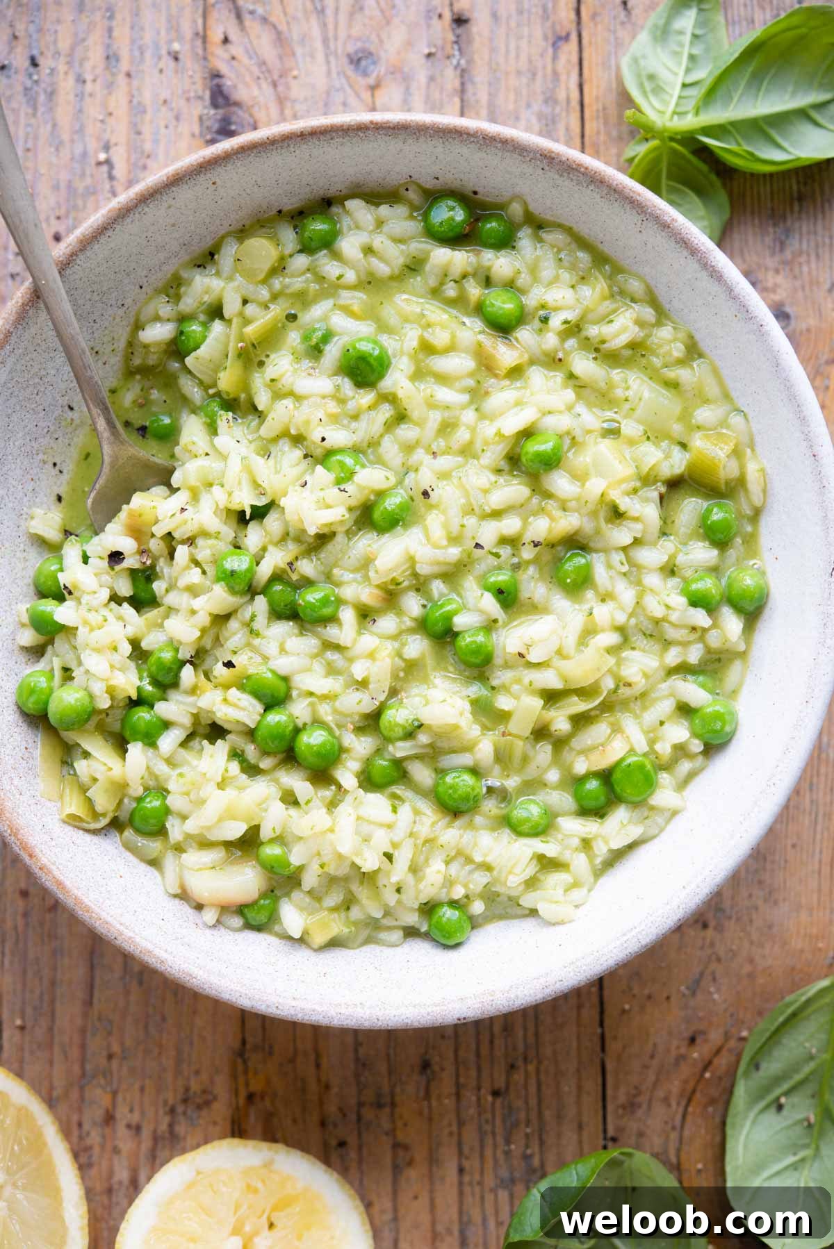 An overhead shot of a bowl of risotto made with peas and pesto on a wooden background with basil and lemons at the side
