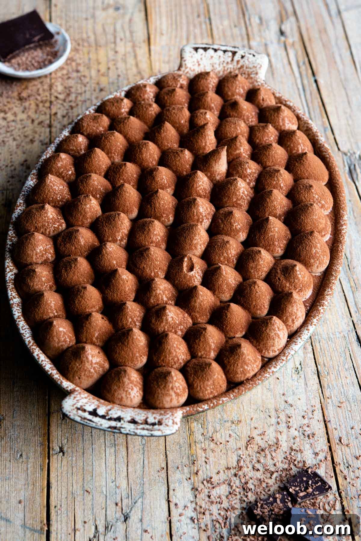 Chocolate tiramisu in an oval dish sitting on a rustic wooden background with pieces of chocolate at the side