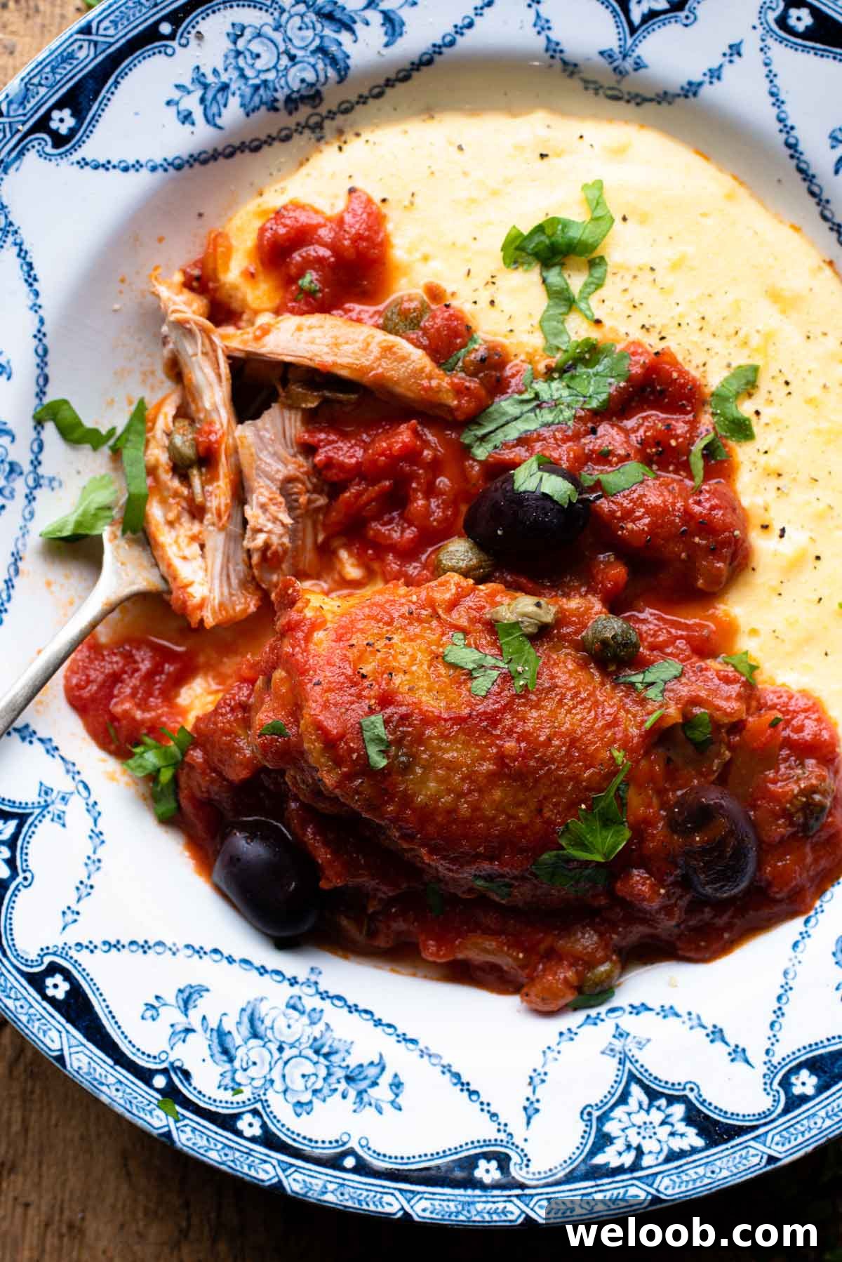 Chicken in Puttanesca sauce in a bowl with polenta sitting on a wooden surface.