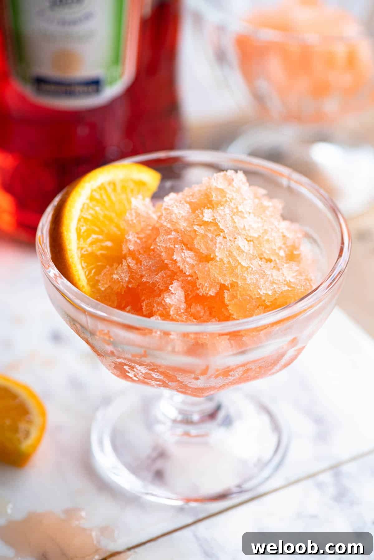 Aperol granita in a small glass bowl with a slice of orange at the side sitting on a marble board.