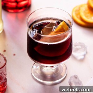 A square image of an Italian red wine spritz on a light work surface with ice and oranges around.
