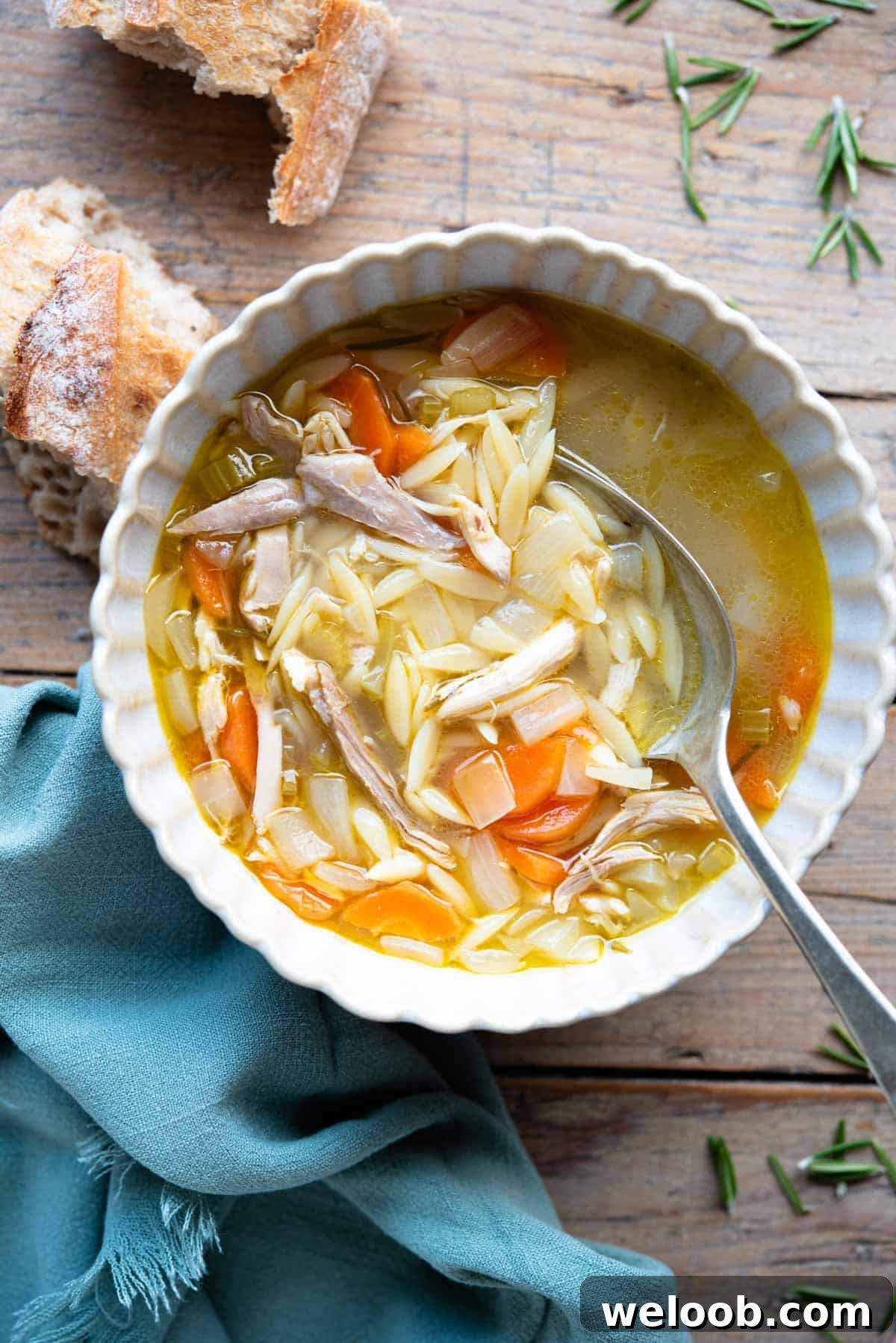 An overhead shot of a bowl of lemon chicken orzo soup on a wooden surface with crusty bread at the side.
