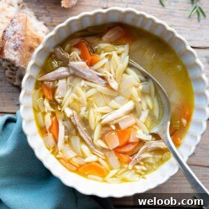 An overhead shot of lemon chicken orzo soup on a bowl. The background is wooden with crusty bread at the side.