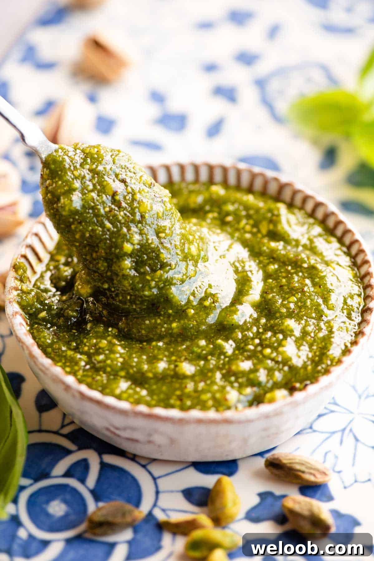 A close up of a spoon in a dish of pistachio pesto sitting on a blue patterned background with pistachios around.