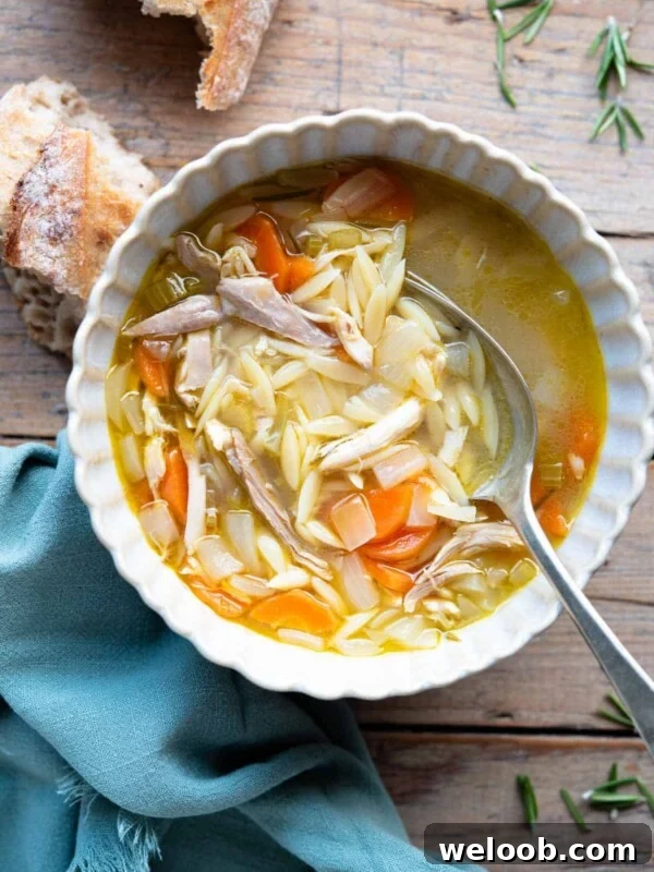 An overhead shot of a bowl of lemon chicken orzo soup on a wooden surface with crusty bread at the side.