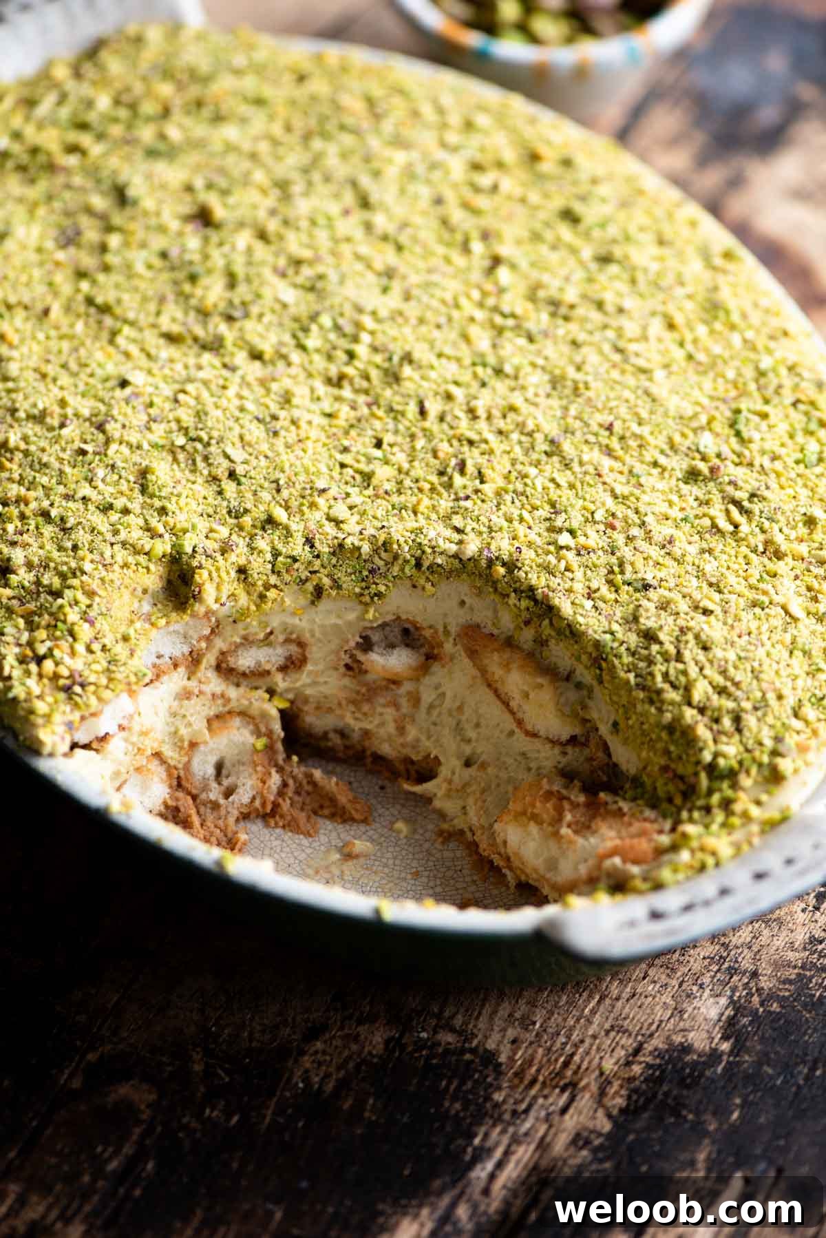 A side shot of a pistachio tiramisu in an oval dish with a slice removed. The background is wooden.
