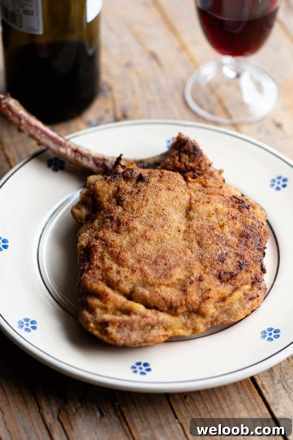 A breaded veal chop sitting on a plate with a glass and bottle of red wine in the background.