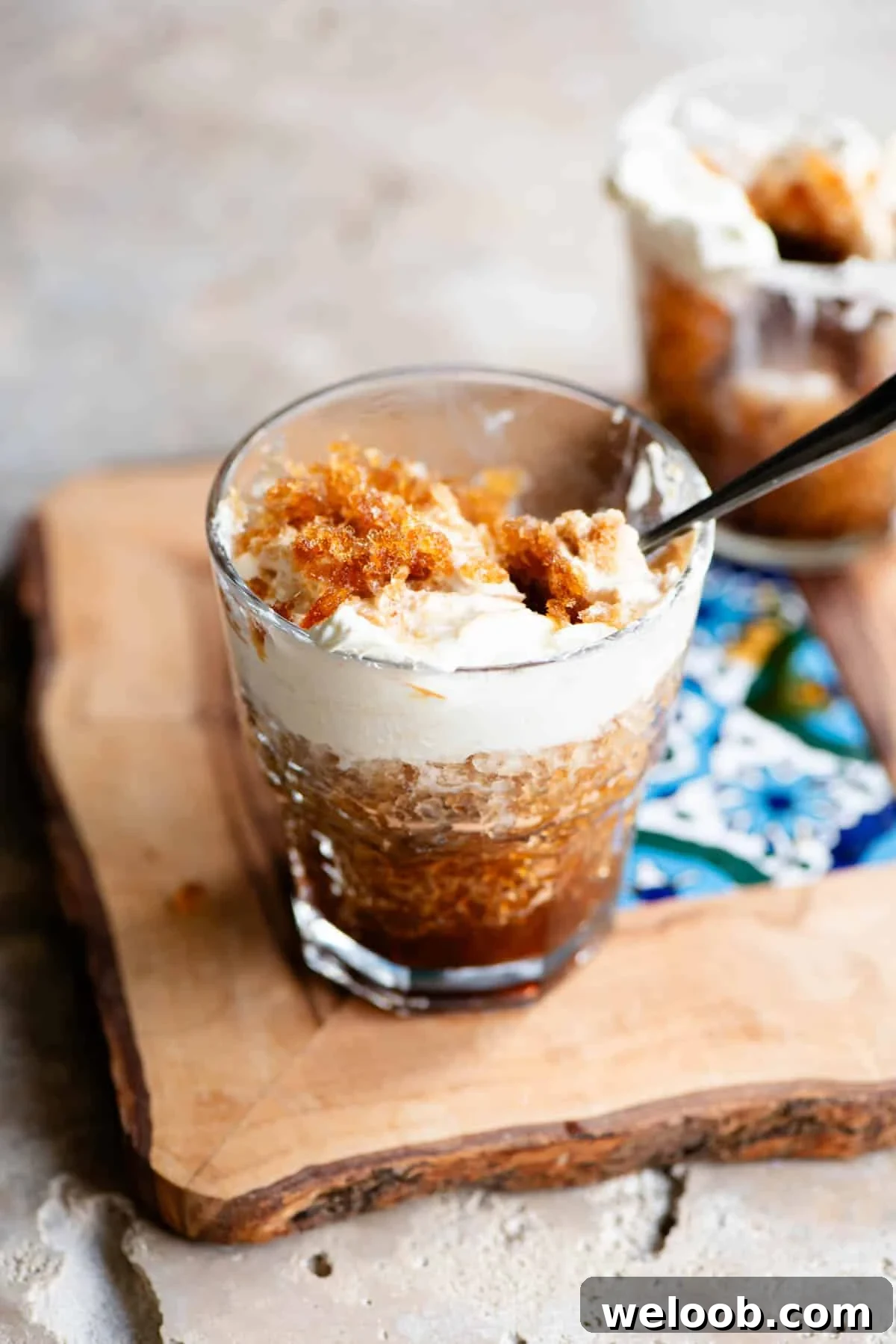 A glass filled with coffee granita and whipped cream sitting on a wooden serving board with a patterned tile in the middle.