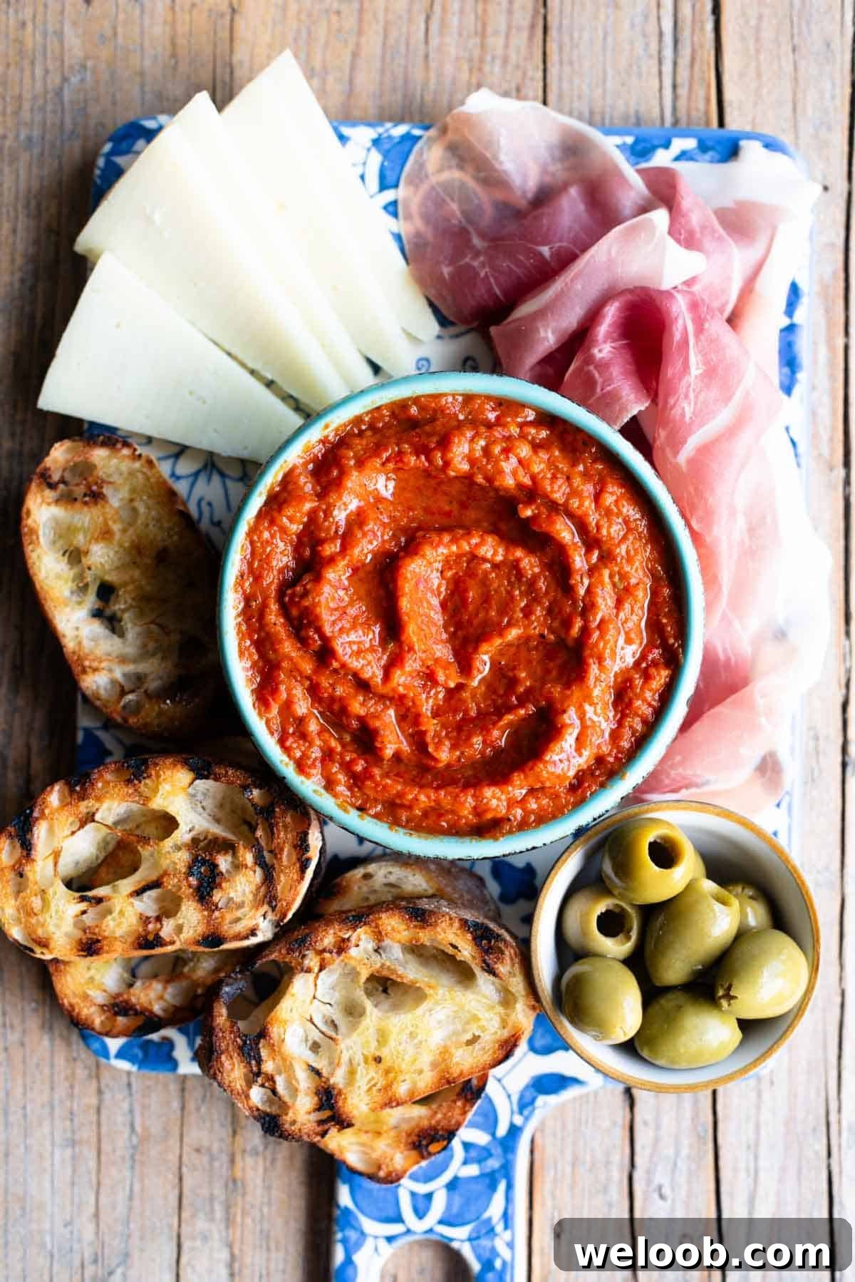 An overhead shot of Ajvar spread in a blue bowl sitting on a serving board with cured meats, cheese and olives.