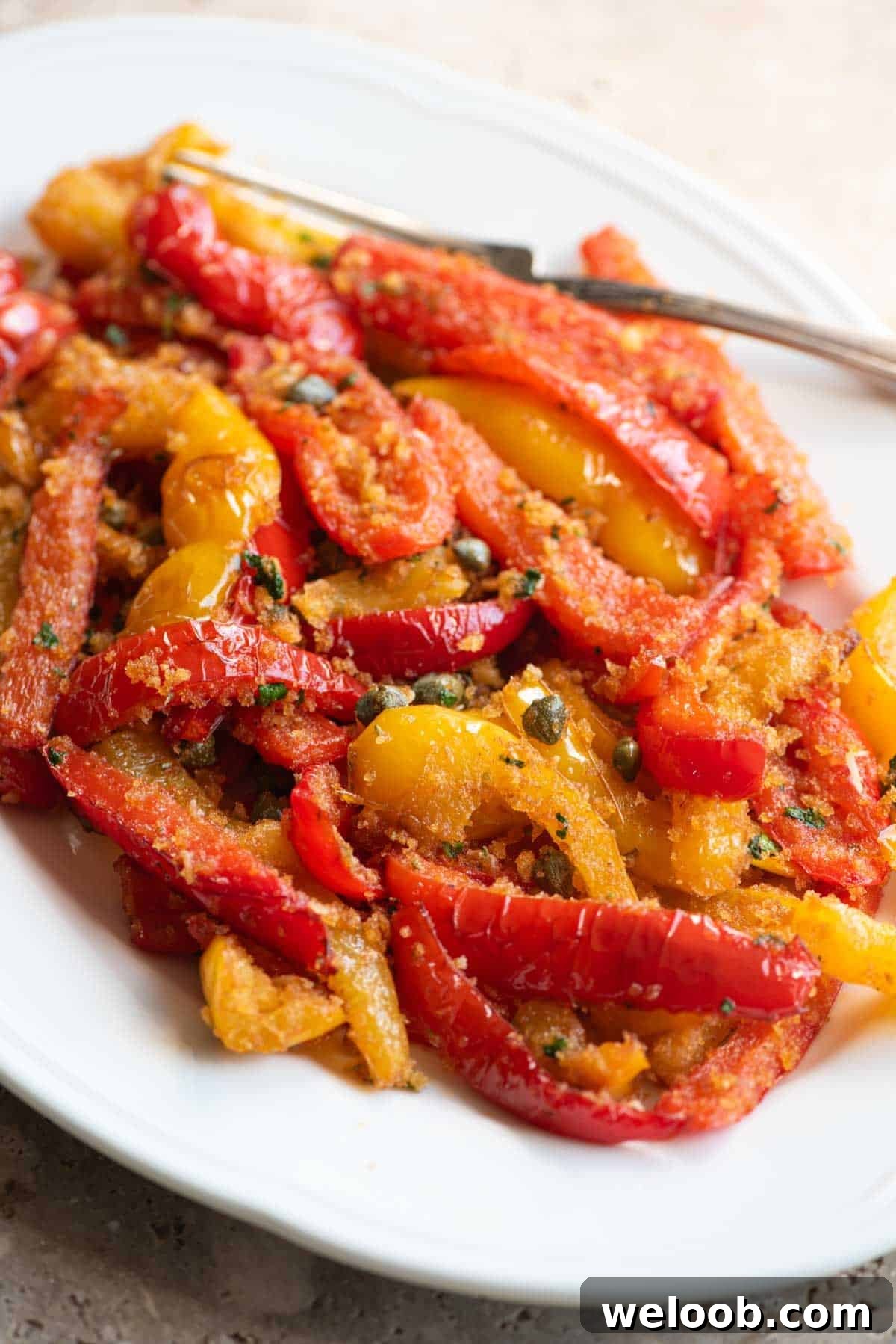 A close up side shot of mixed red and yellow peppers on a white servings dish with capers and breadcrumbs.
