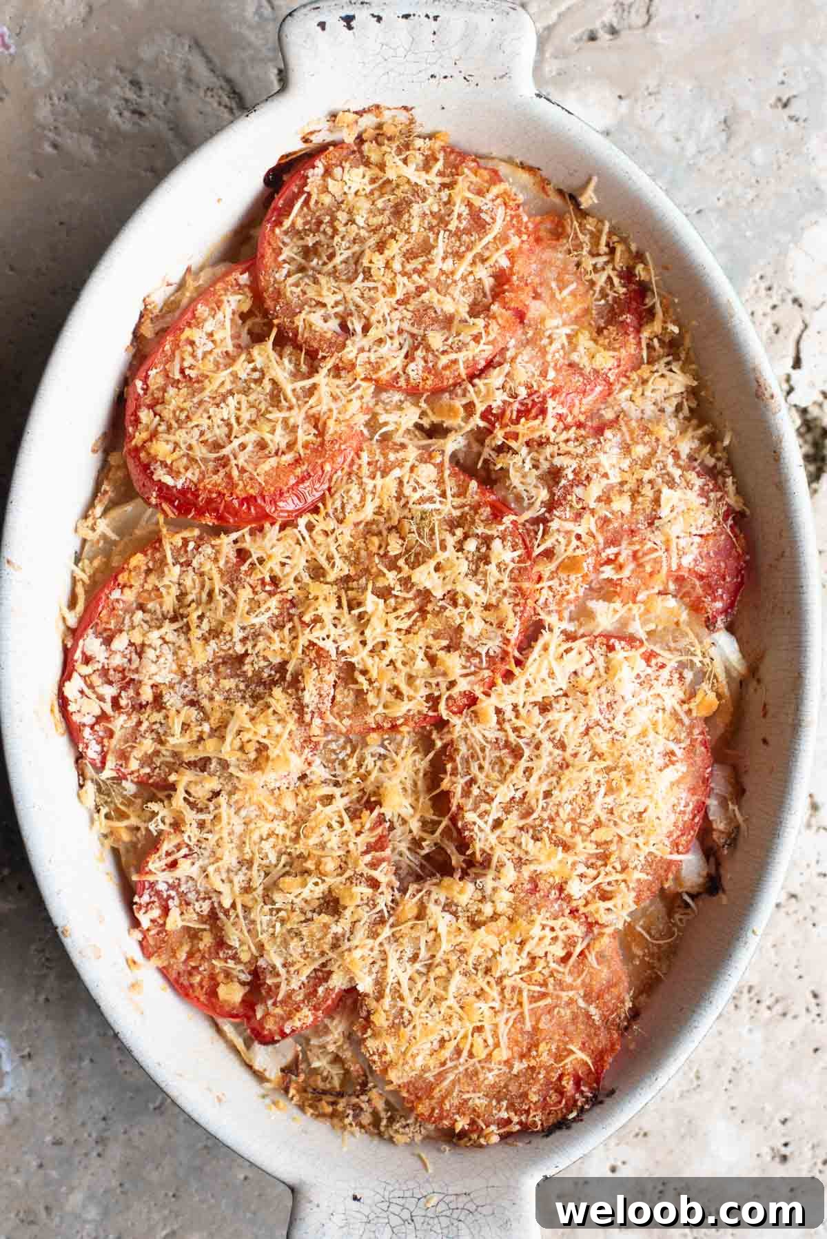 An overhead shot of an Italian potato and tomato gratin in an oval dish sitting on a Travertine background.
