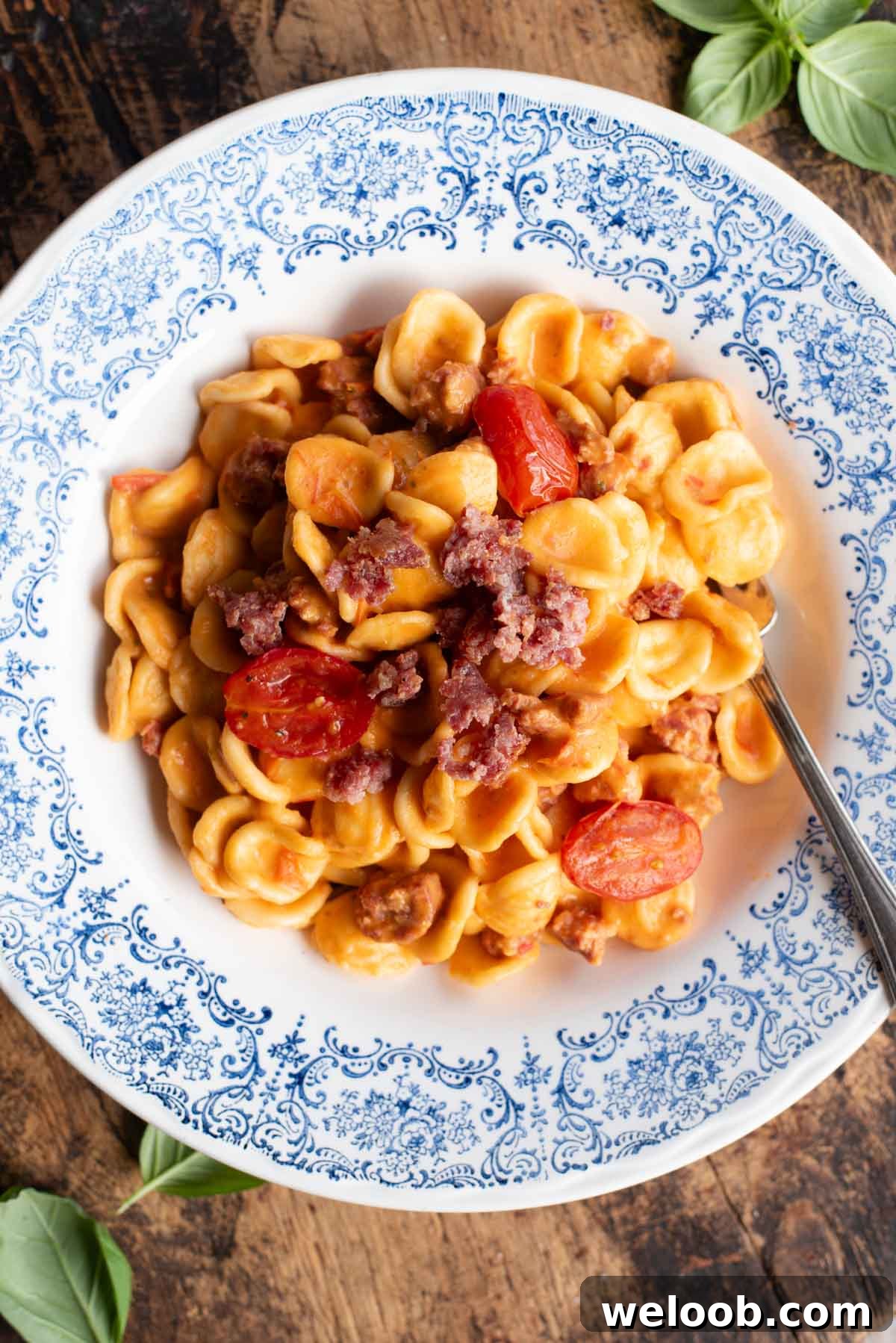 An overhead shot of pasta with sausage, tomato and burrata in a blue and white bowl.