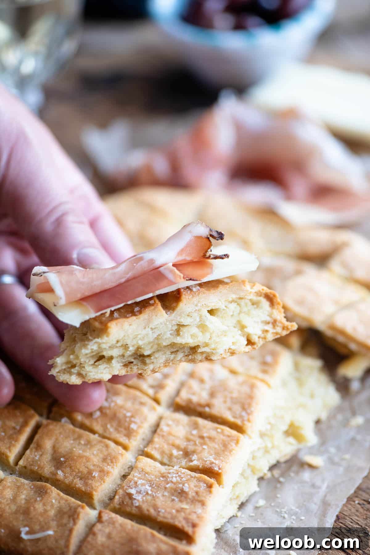 A close up of a hand holding a piece if Pizza Scima with cured meat.