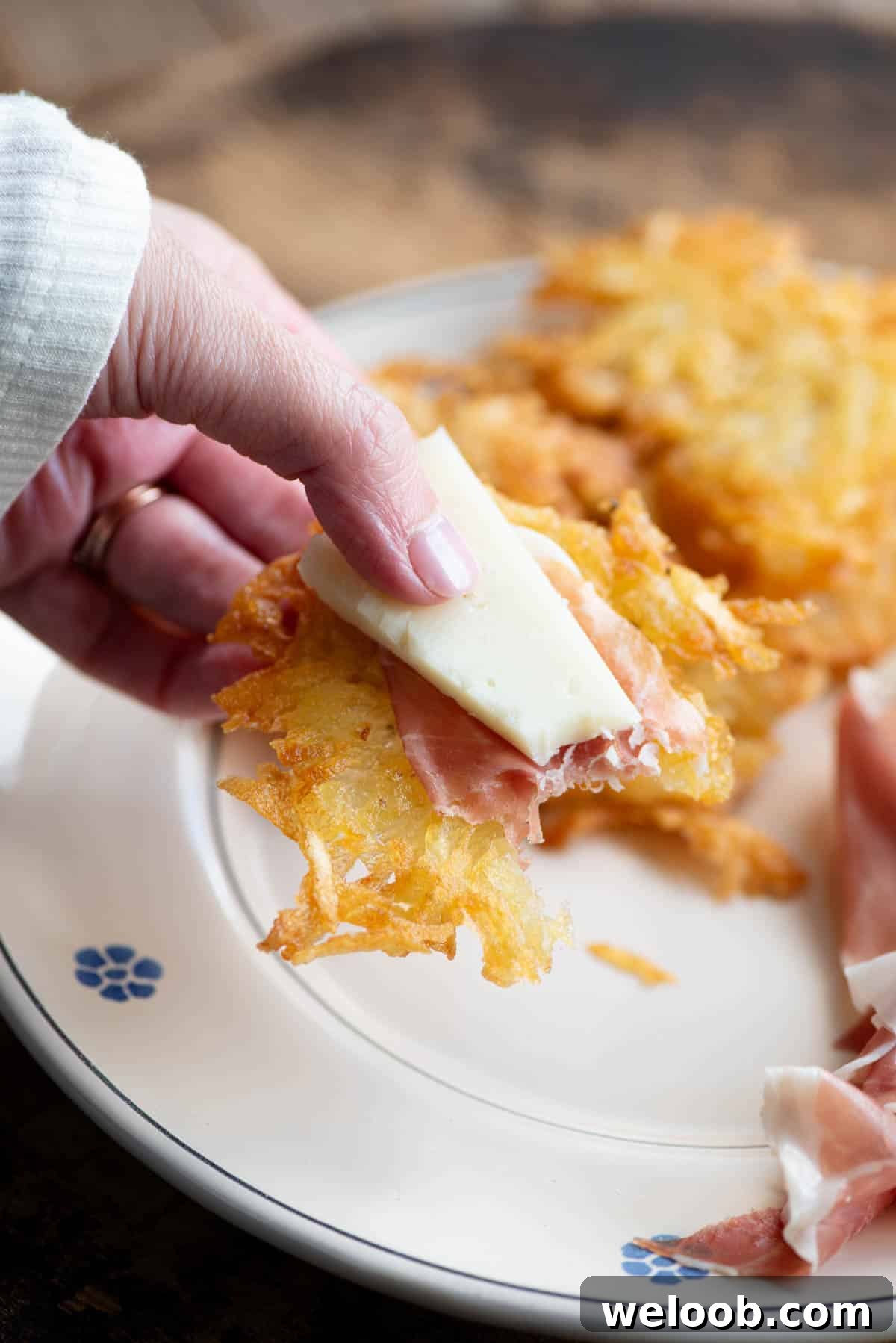 A close up of a hand holding a potato fritter topped with cured meat and cheese.