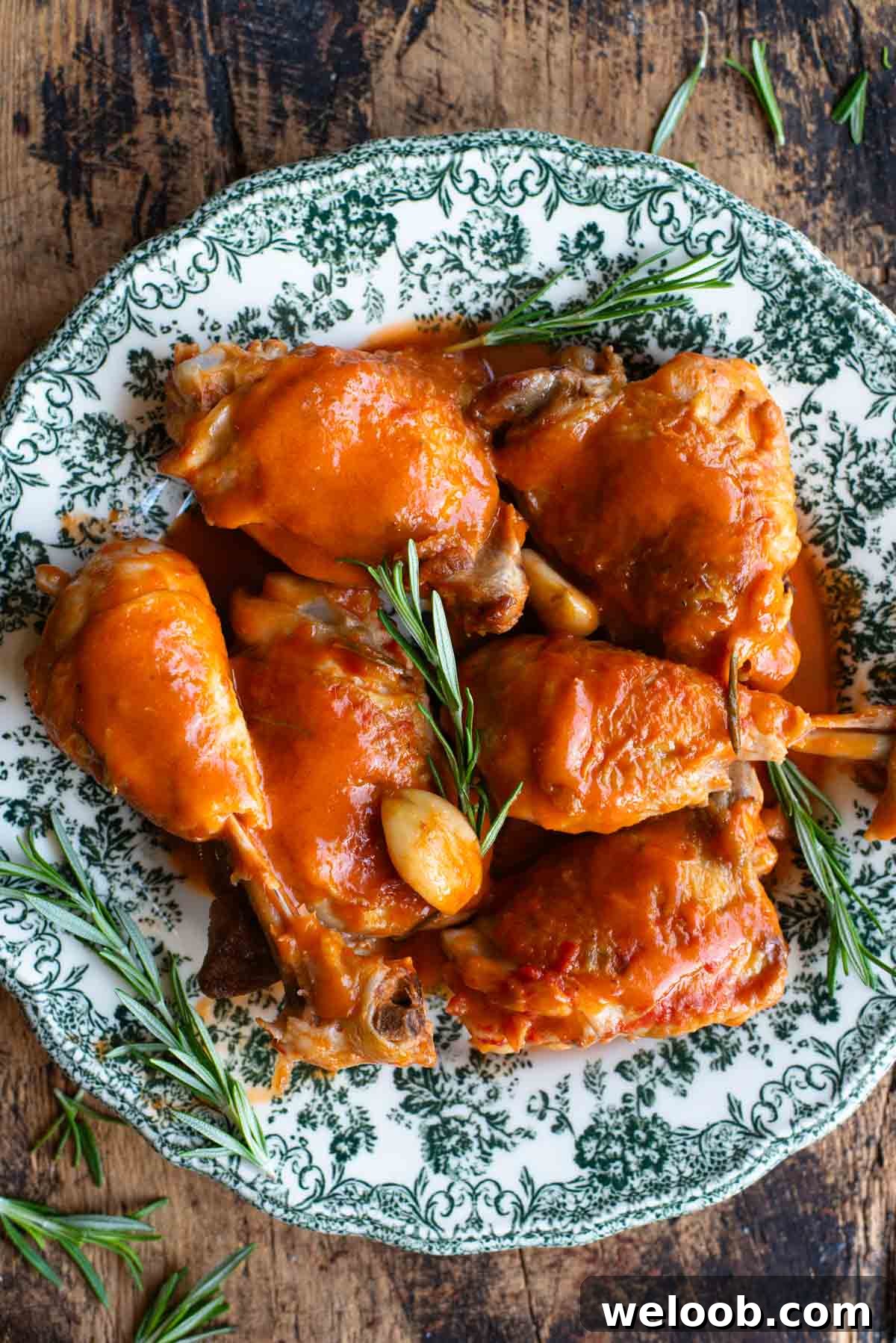 An overhead shot of chicken thighs and drumsticks on a green patterned plate with rosemary and garlic.
