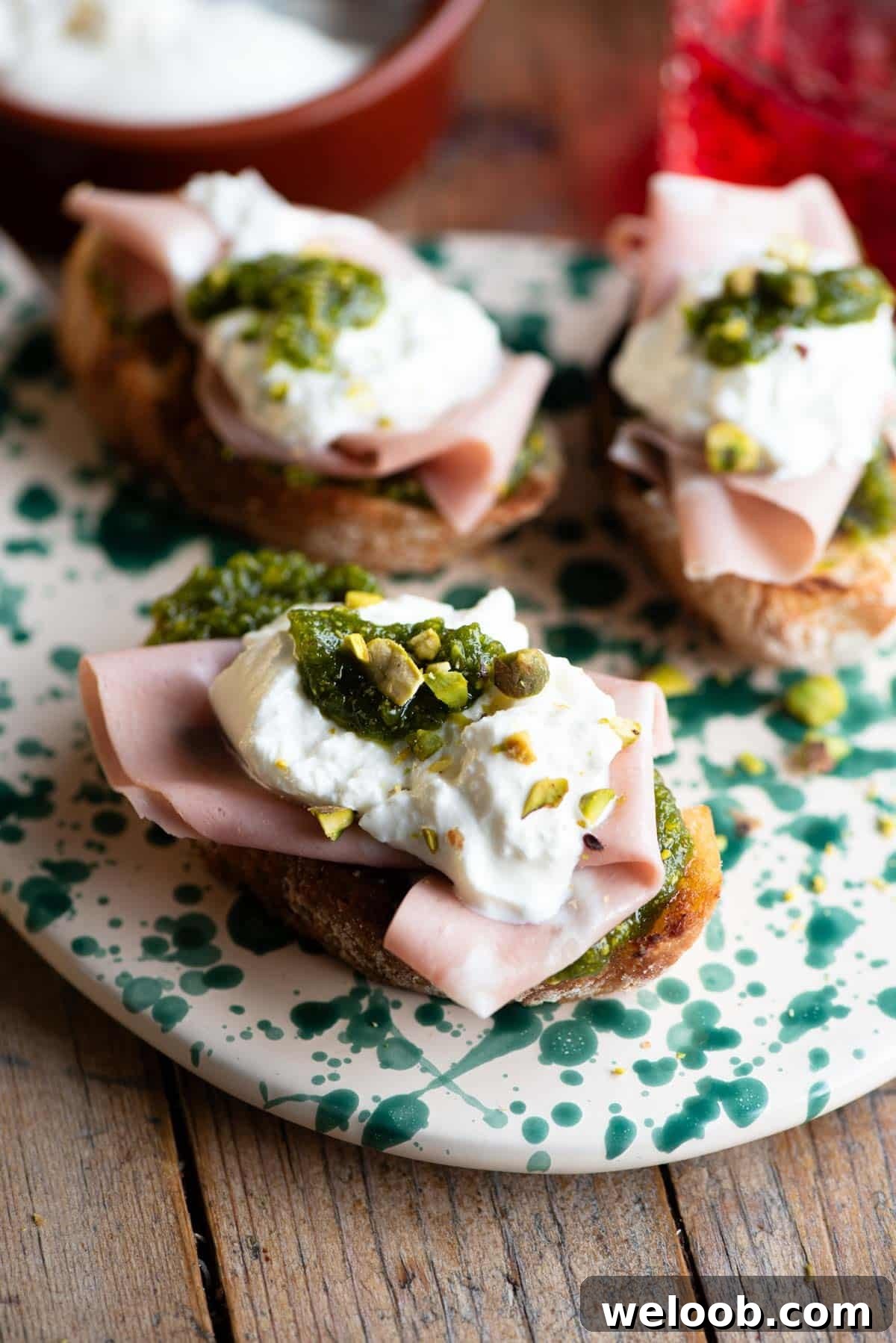 A close up of three crostini on a spotted serving board topped with mortadella, burrata and pistachios.