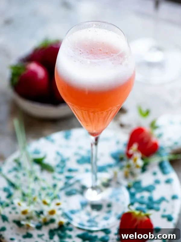 A close up of a strawberry bellini (rossini) cocktail sitting on travertine with strawberries and flowers around.