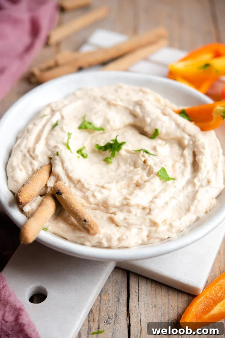 A side shot of a white bean dip with breadsticks and bell peppers around it