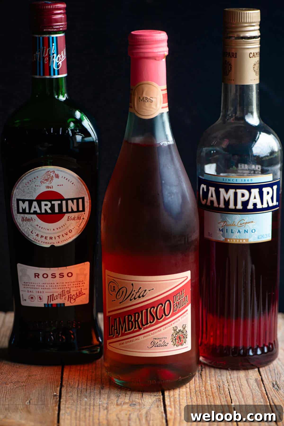 A bottle of Martini rosso, Lambrusco and Campari sitting on a wooden table with a black background.