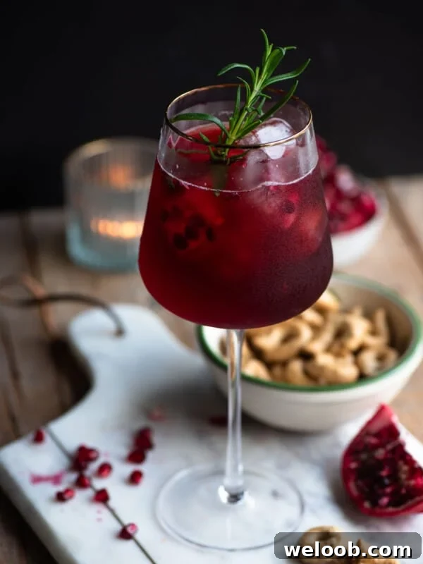 A pomegranate spritz in a tall wine glass with a gold rim and a sprig of rosemary inside.