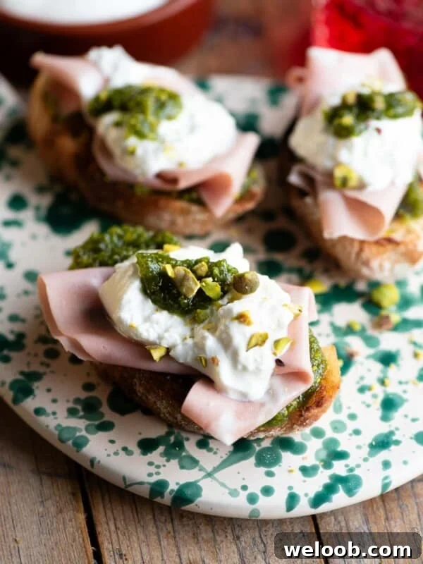 A close up of three crostini on a spotted serving board topped with mortadella, burrata and pistachios.