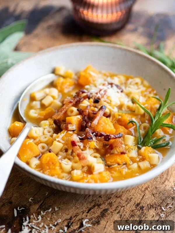 A butternut squash and pasta soup topped with pancetta and a sprig of rosemary. The bowl is on a wooden table.