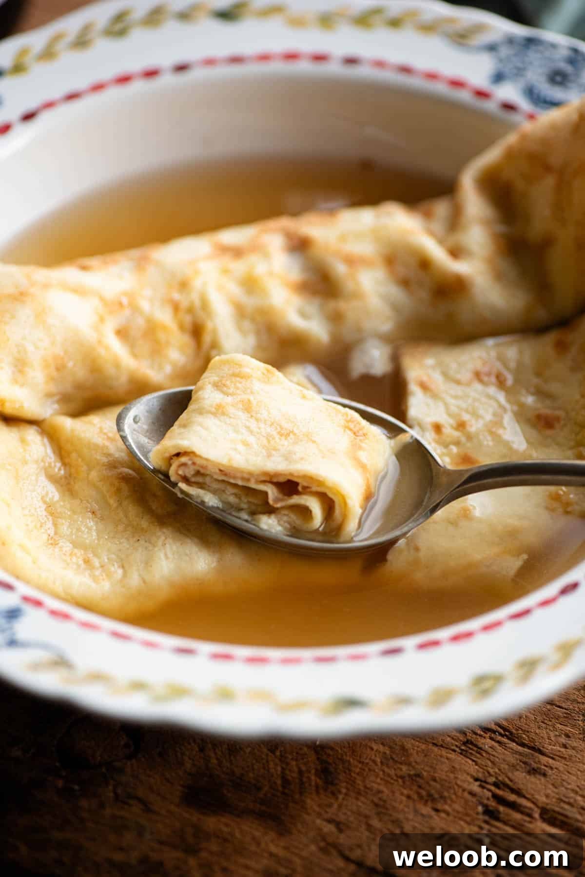 A close up of a spoon in a bowl of broth picking up a spoonful of crepe.