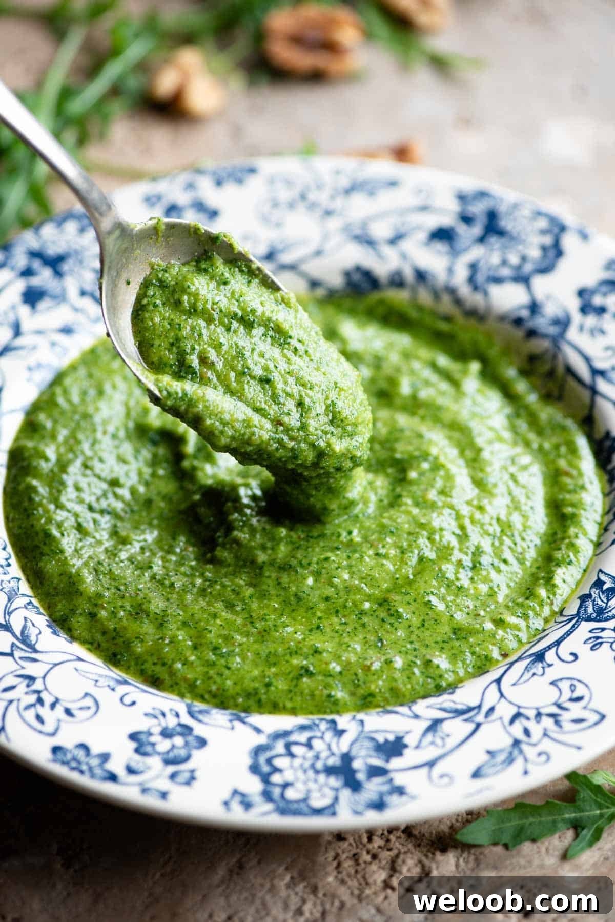 A close up of a spoon picking up some arugula pesto from a blue and white bowl.