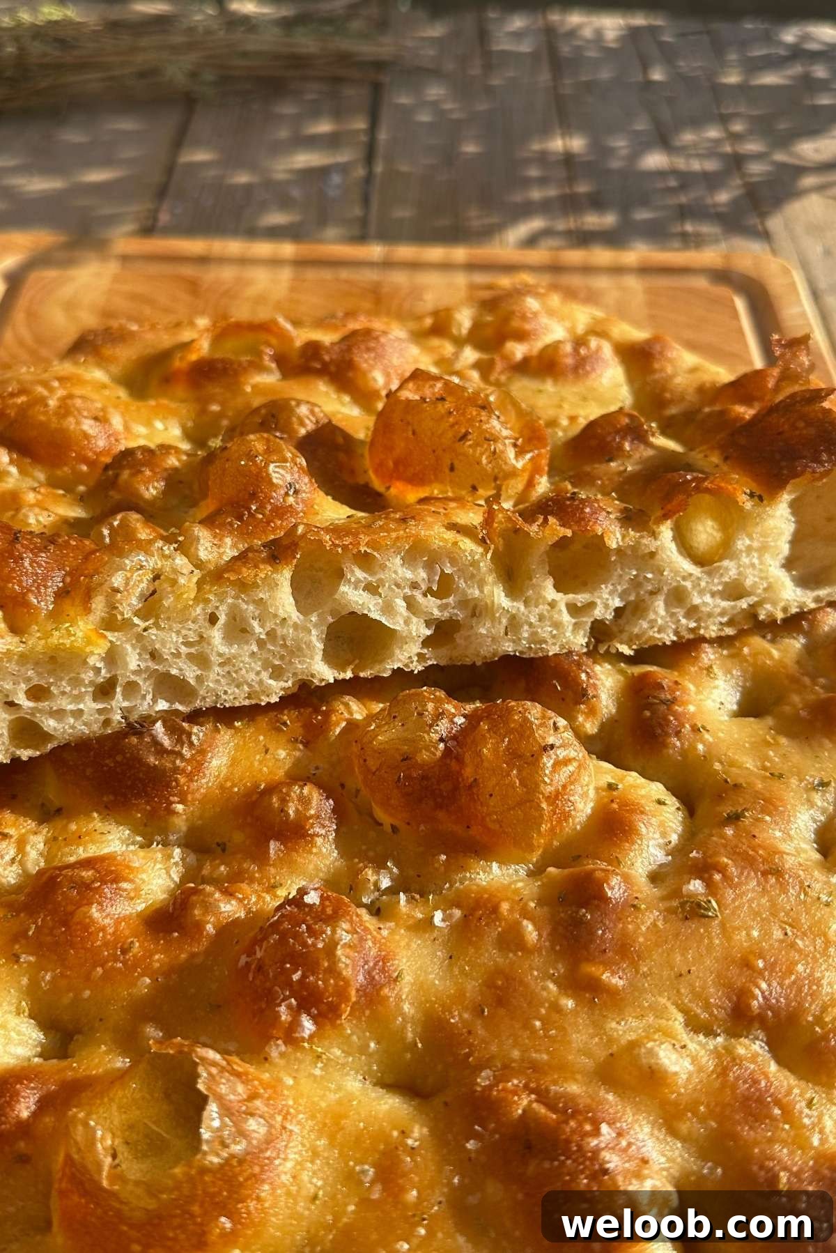A golden and airy focaccia cut in half to show it's bubbly texture sitting on a wooden chopping board.