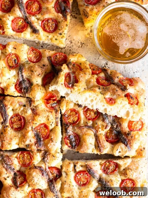 An overhead shot of a cherry tomato and anchovy focaccia cut into slices with a glass of beer at the side.