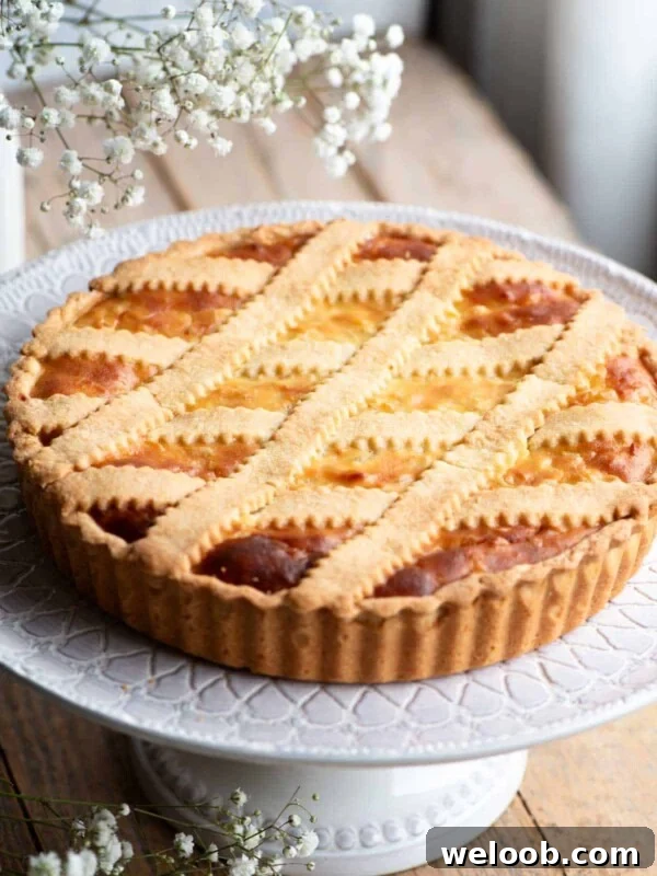 Pastiera Napoletana on a cake stand sitting on a wooden surface with white flowers at the side.