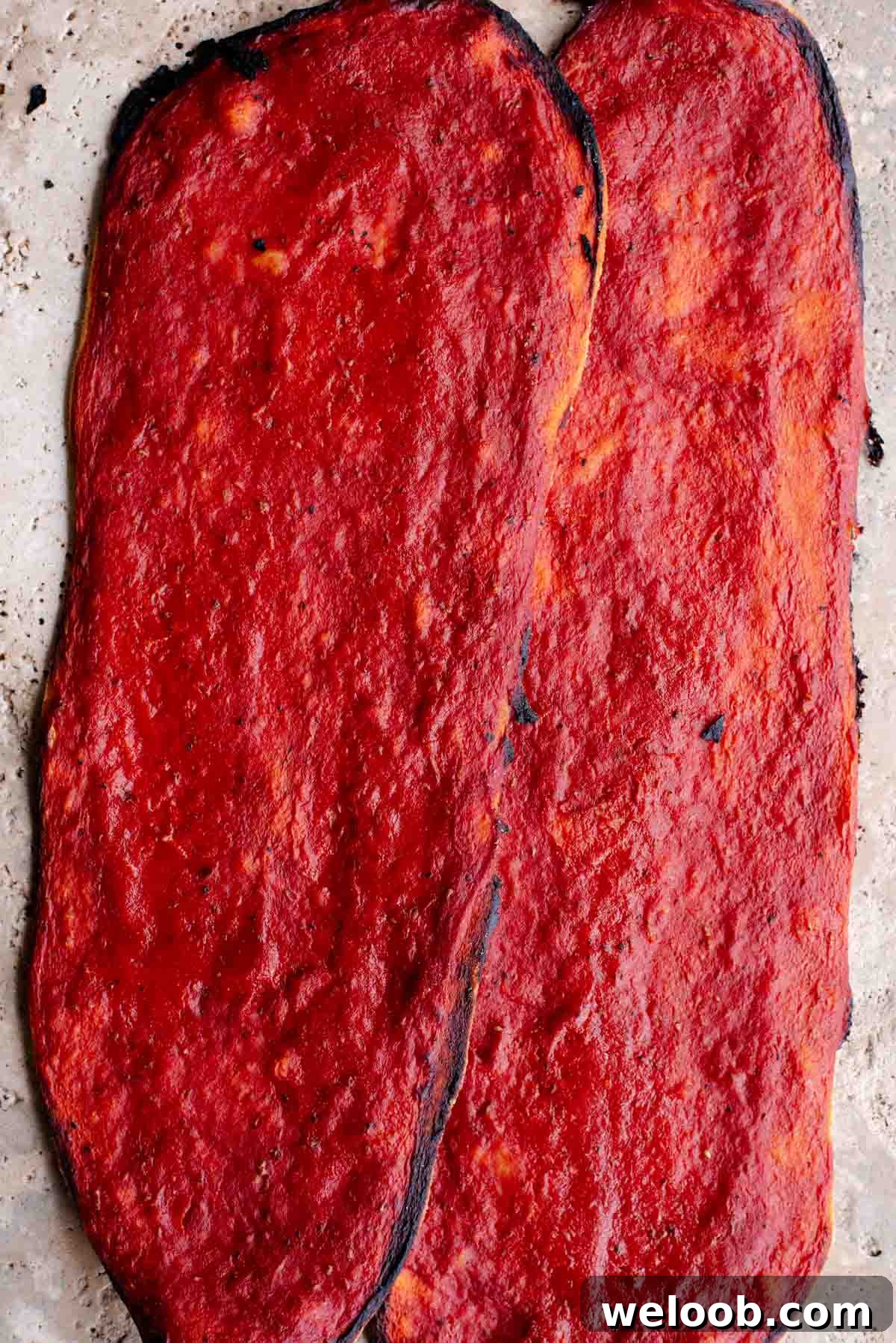 Two long thin and crispy pizzas covered in tomato sauce sitting on a travertine surface.