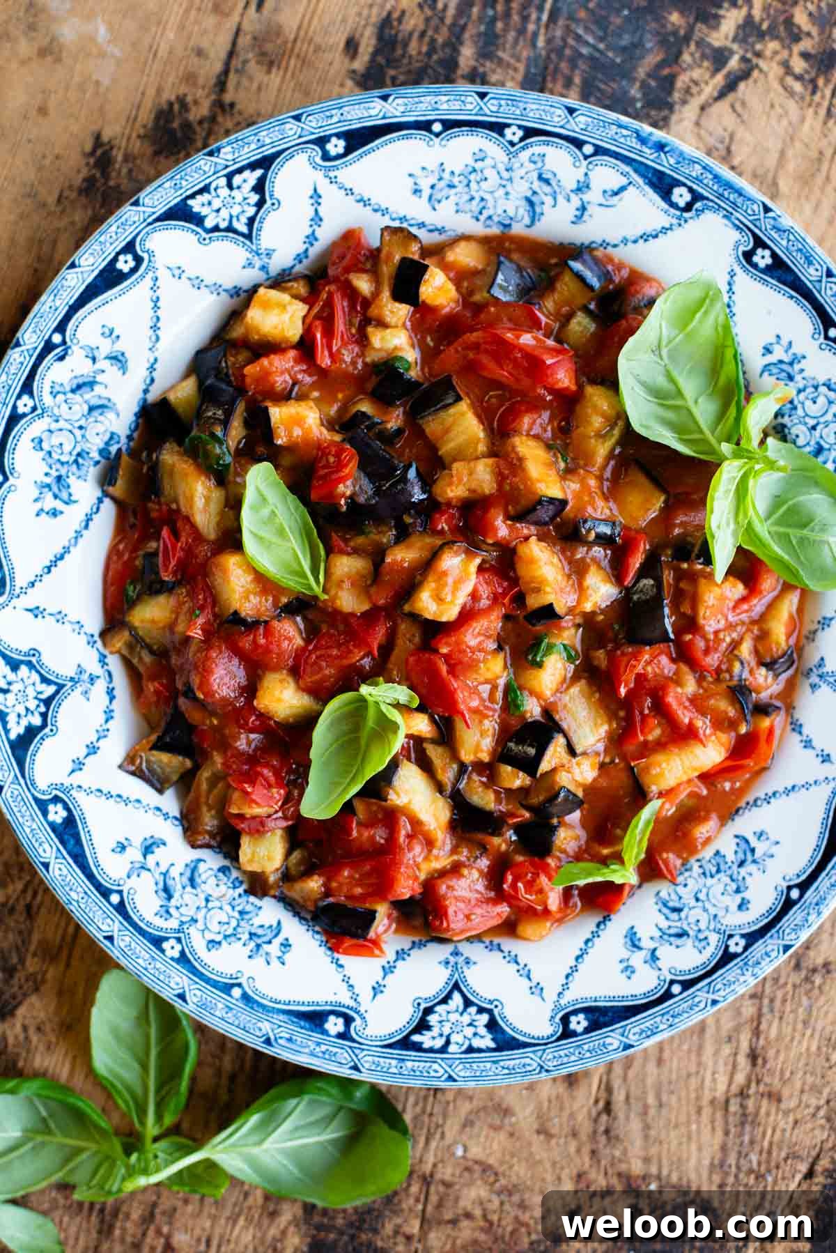 Fried eggplant in tomato sauce with fresh basil in a blue and white bowl on a wooden surface.