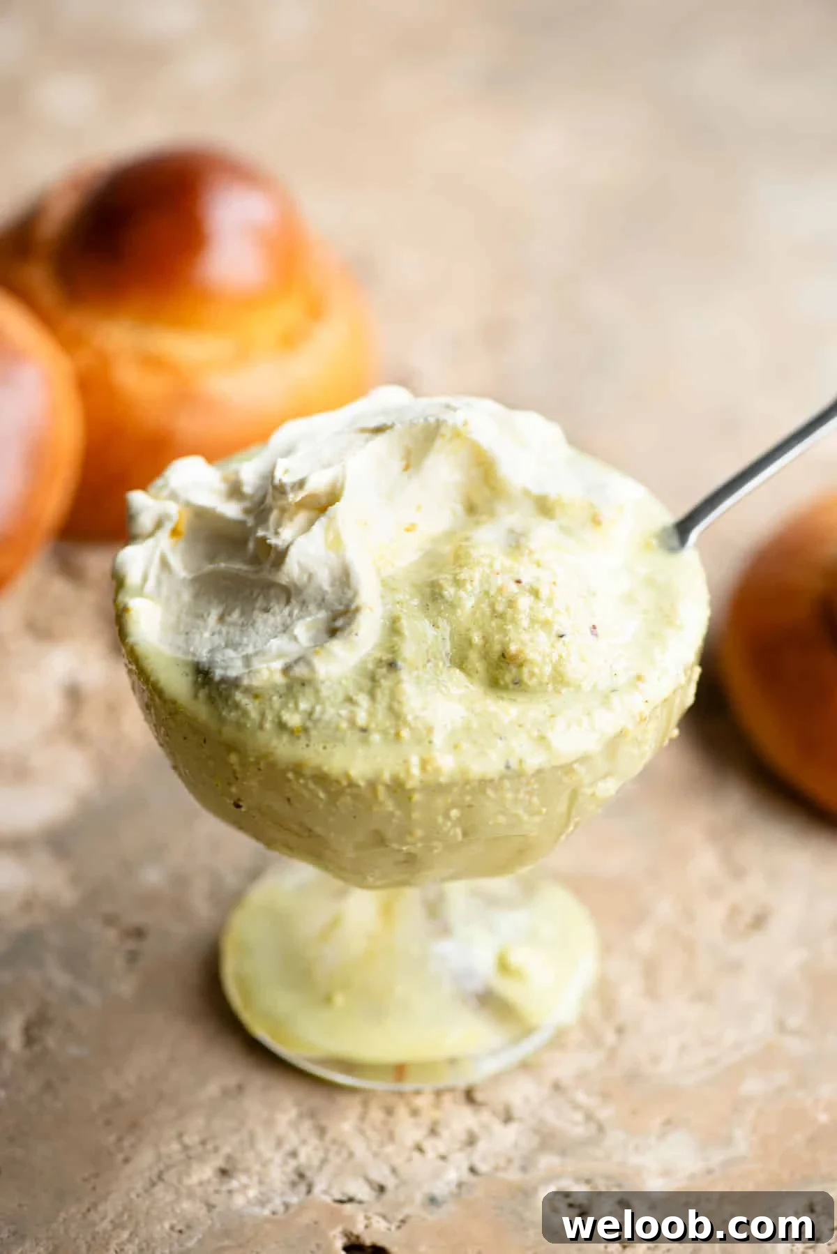 A close up of pistachio granita in a glass serving bowl topped with whipped cream and a brioche bun behind it.