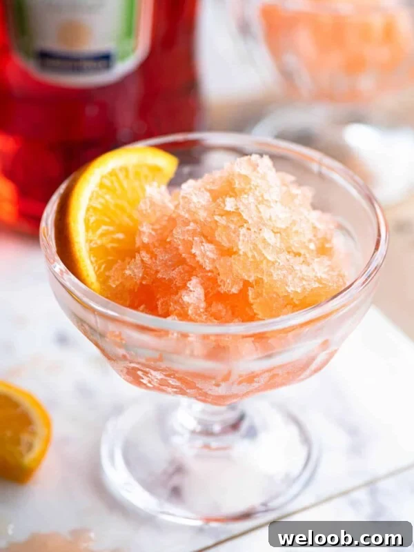 Aperol granita in a small glass bowl with a slice of orange at the side sitting on a marble board.
