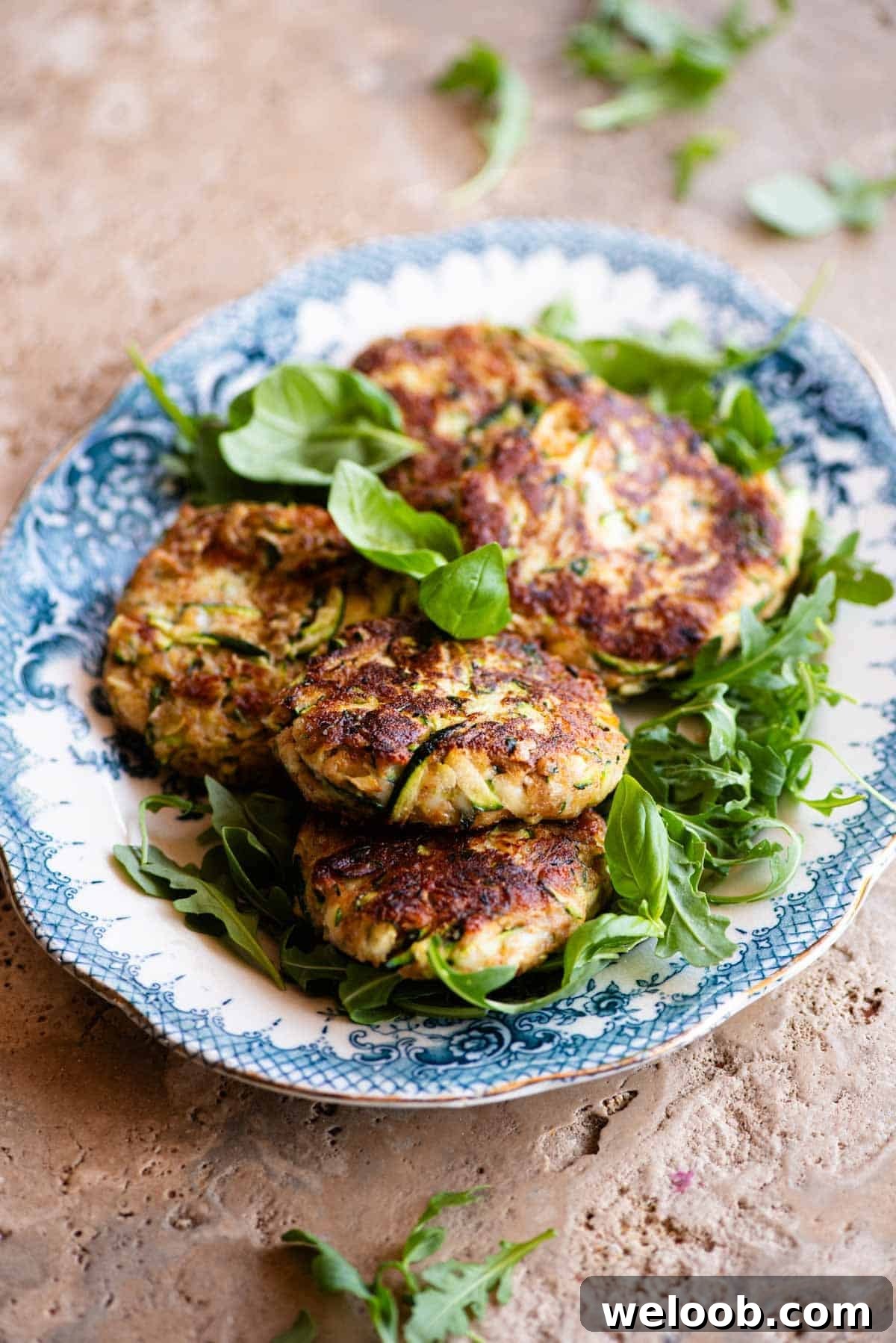 Five zucchini cakes (patties) sitting on top of arugula on a blue and white vintage style serving platter sitting on a travertine surface.