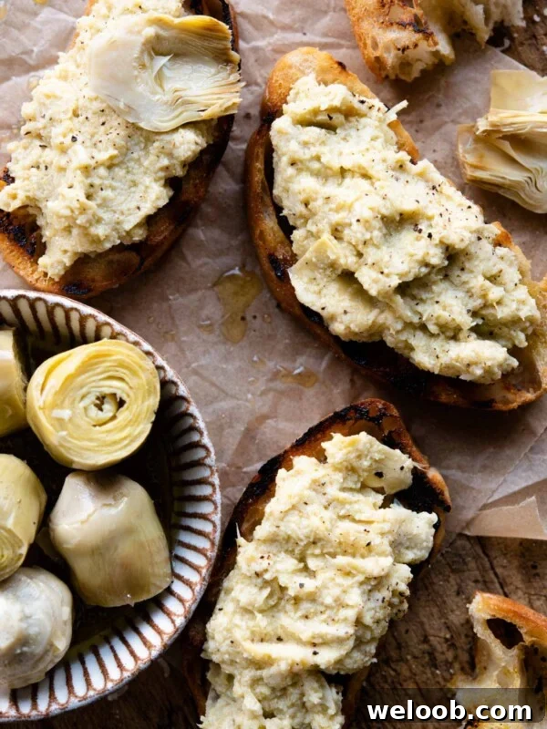 Three slices of artichoke bruschetta sitting on a wooden serving board with a small ramekin of artichokes beside it.