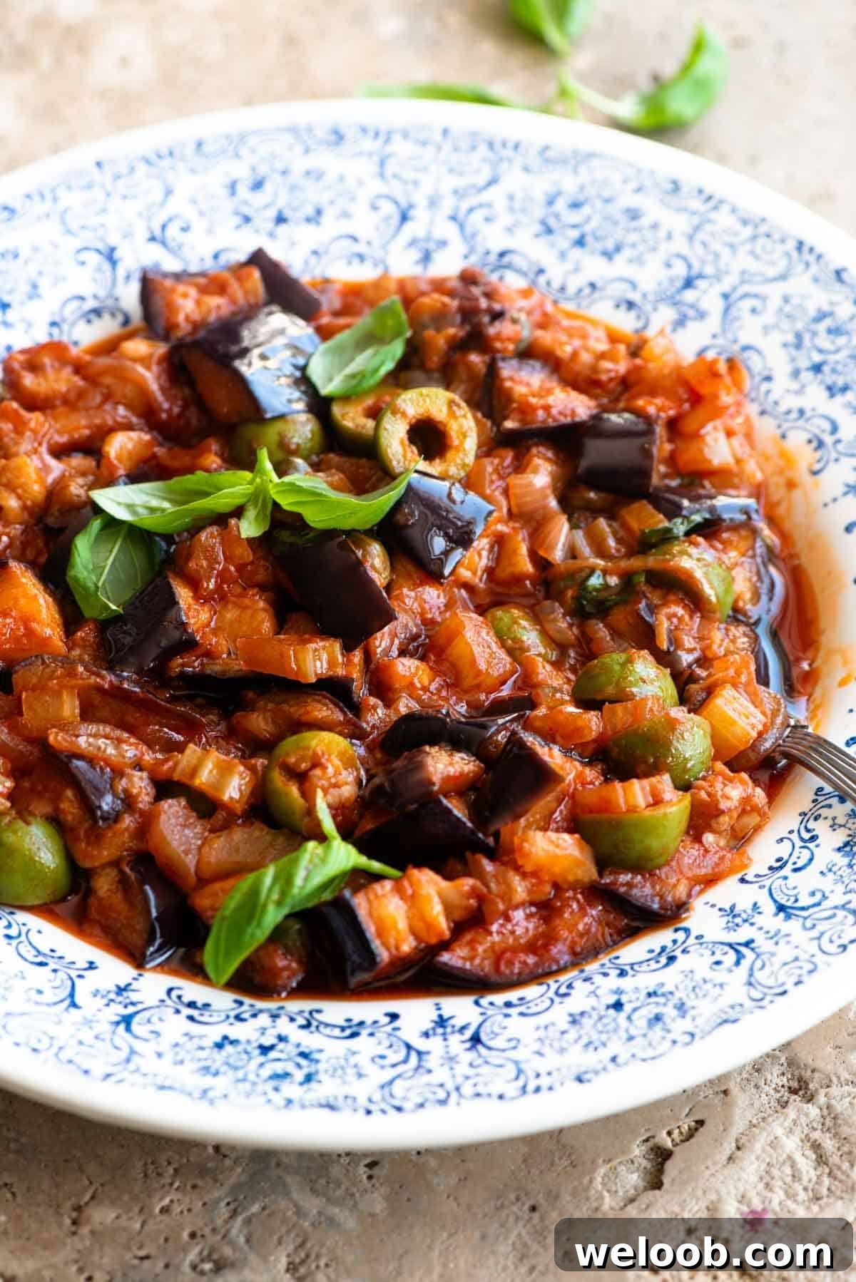 A side shot of caponata in a blue and white bowl topped with fresh basil leaves.