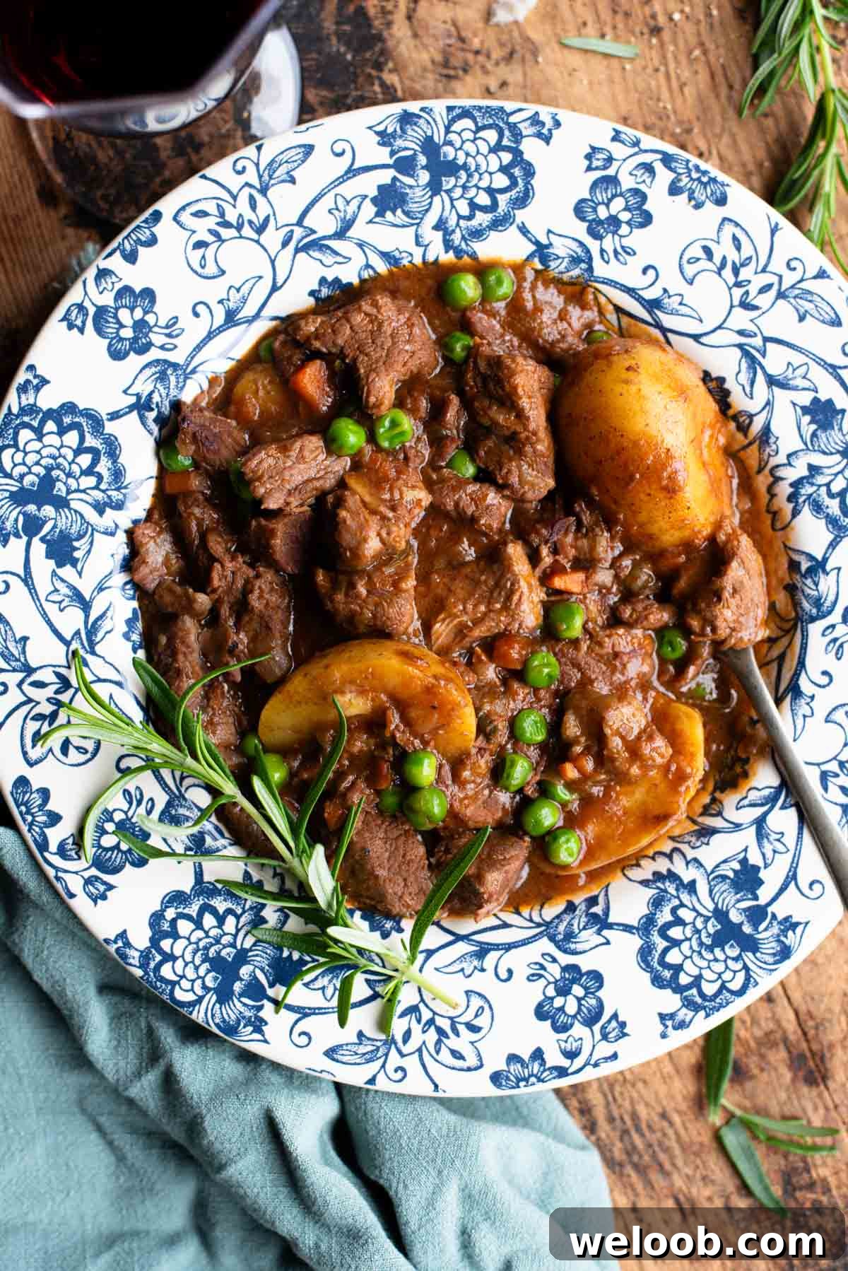 Italian beef stew with potatoes and peas in a blue and white floral bowl with rosemary and red wine at the side.