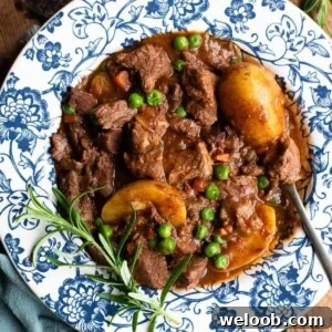 A close up of a beef stew with potatoes and peas in a blue and white bowl with rosemary.