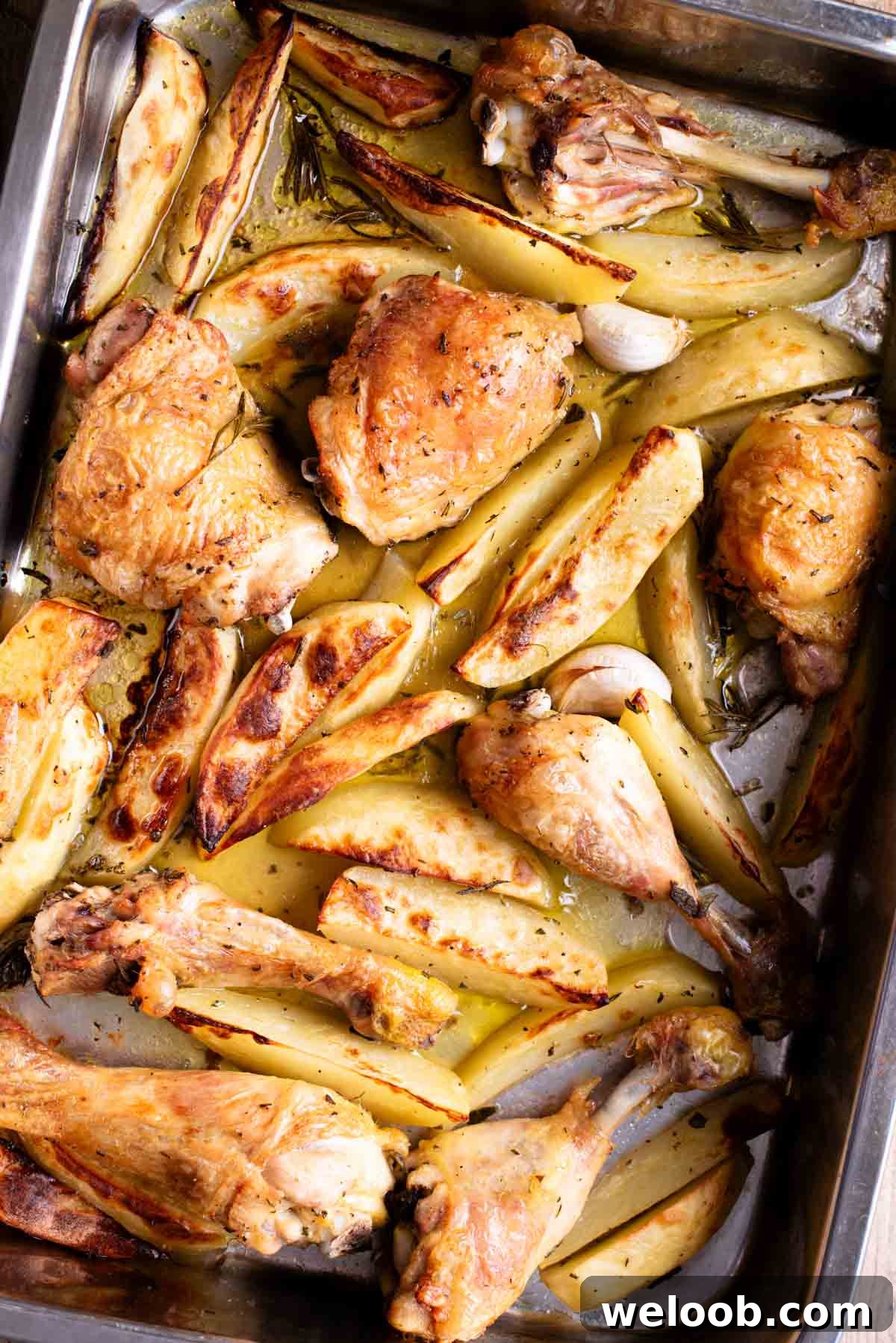 A close up overhead shot of chicken and potato wedges in a roasting tray with garlic and rosemary.