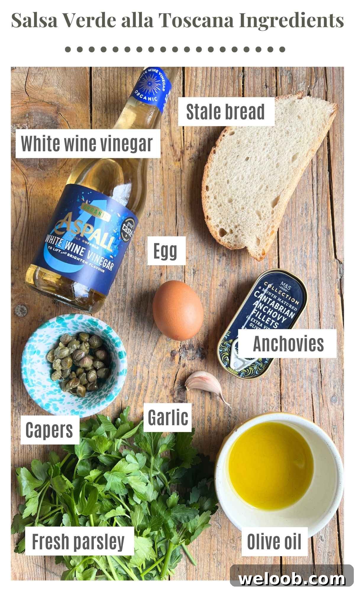 All the ingredients needed to make a Tuscan salsa verde on a wooden board. The text labels read; white wine vinegar, stale bread, garlic, anchovies, egg, capers, parsley, olive oil.