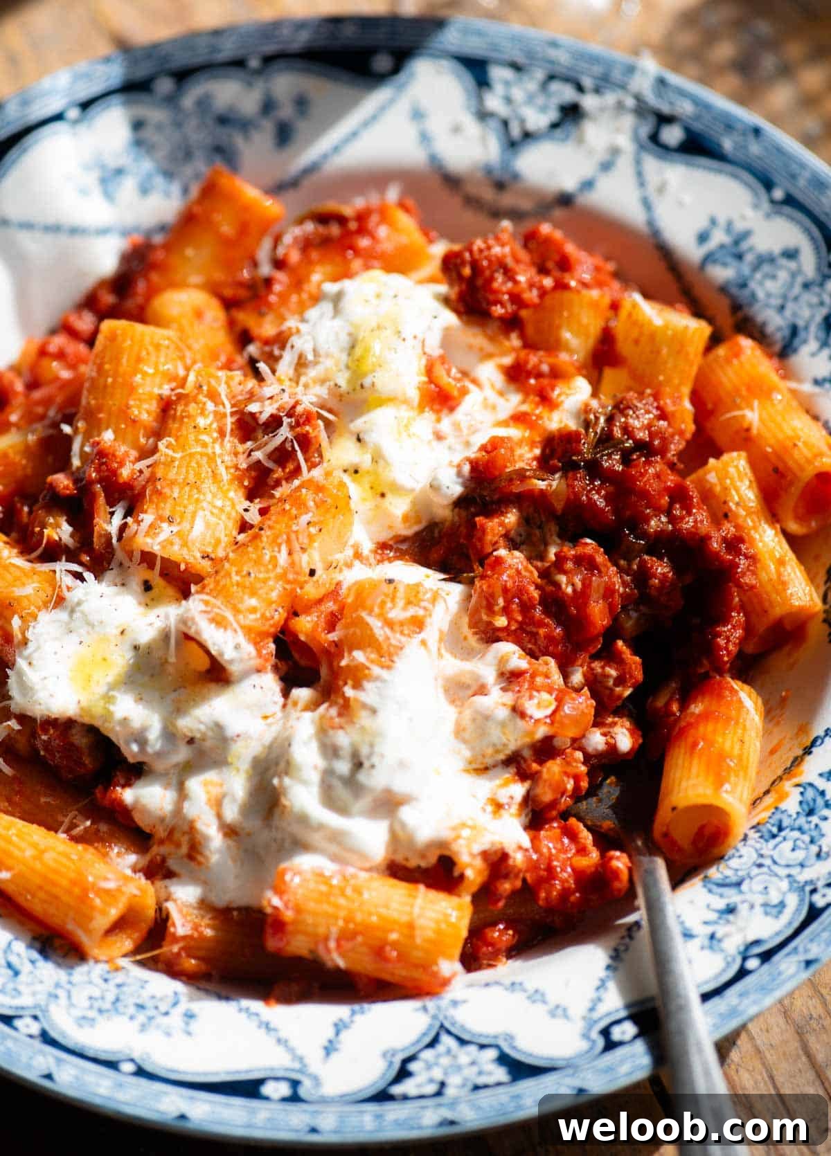 A close up of a spicy sausage ragu topped with stracciatella cheese in a blue and white bowl and a fork at the side.