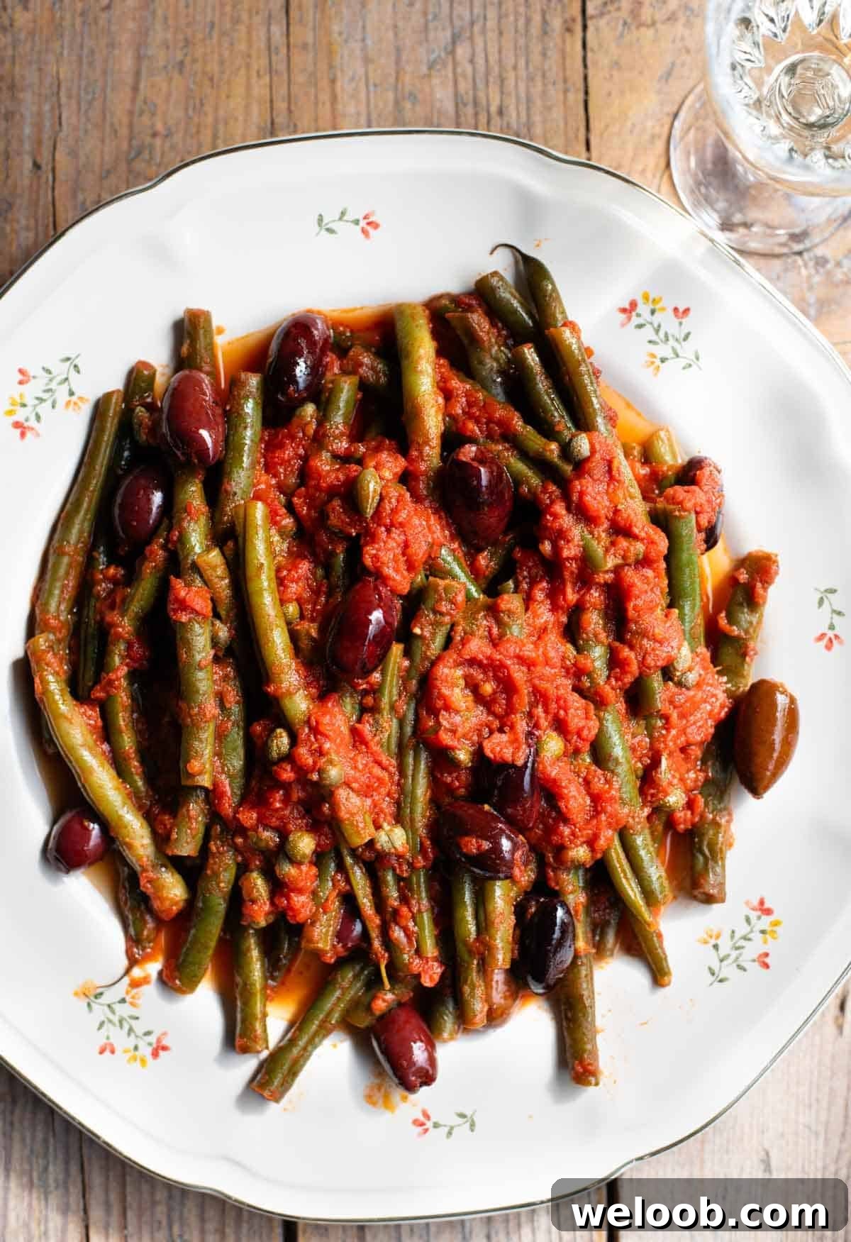 A close up of green beans in tomato sauce with olives and capers on a white floral plate and rustic wooden background.