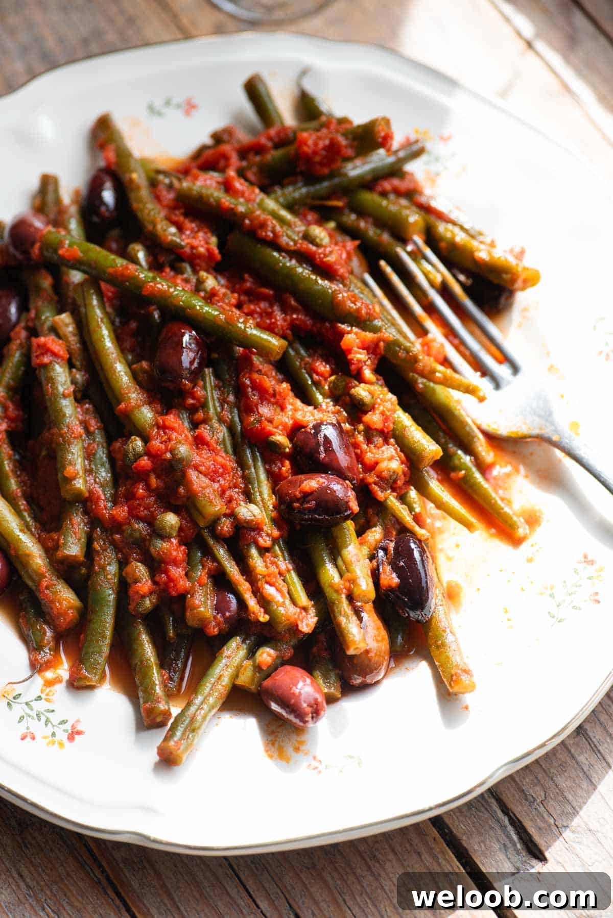 A side shot of green beans with tomatoes, olives and capers on a white plate with a fork at the side.