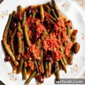 A close up of green beans in tomato sauce with olives and capers on a white plate.