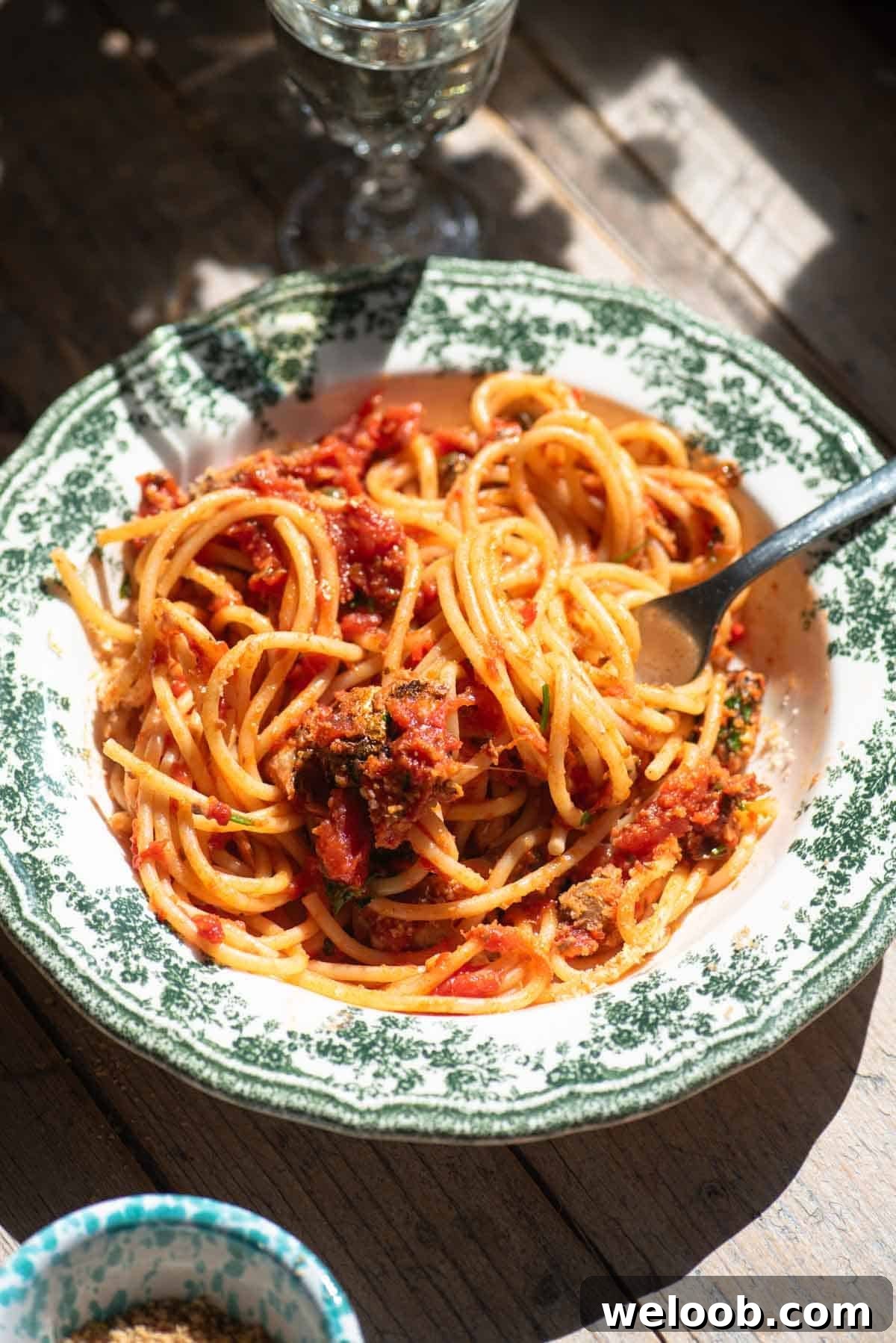 A side shot of tomato sardine spaghetti in a green and white bowl on a rustic wooden surface.