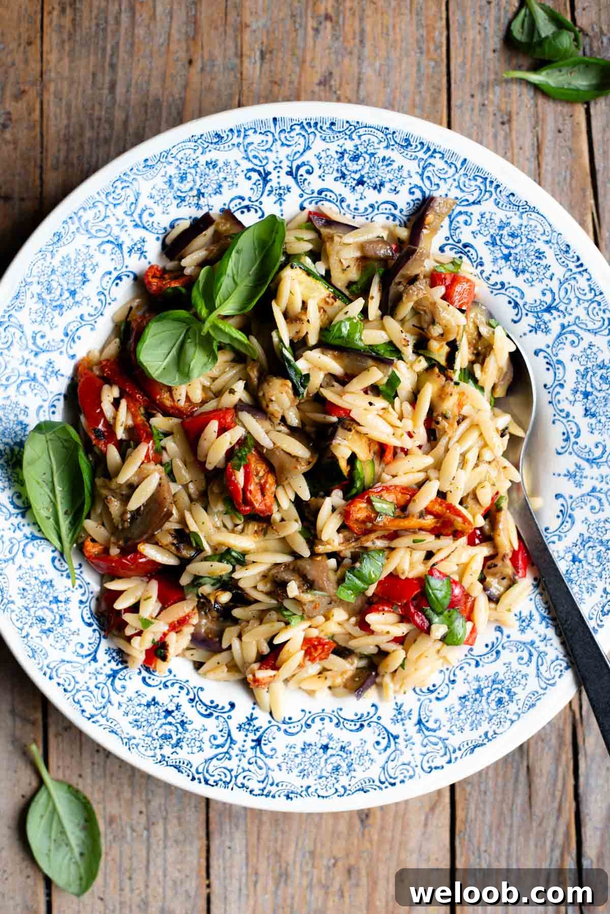 An orzo pasta salad with grilled veg in a blue bowl with a serving spoon at the side. The background is rustic wood.