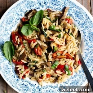A close up of an orzo pasta salad with grilled veg and tomatoes in a blue bowl with basil on top.